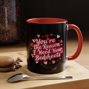 Könnte beinhalten: Schwarze Kaffeetasse mit roter Innenseite und Henkel. Auf der Tasse steht der Text "You're the Reason I Need New Bedsheets", umgeben von Herzen. Ein Löffel und ein Macaron liegen auf einer Holzoberfläche.