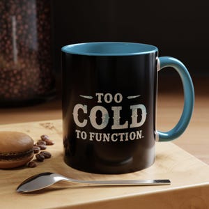 May include: Black coffee mug with a teal interior and handle. The mug features the text "TOO COLD TO FUNCTION" in white. A spoon, macaron, and coffee beans are on a wooden surface.