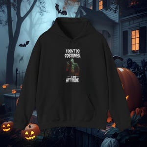May include: Black hooded sweatshirt with a front pocket and drawstring. The graphic on the front features the text "I DON'T DO COSTUMES, I DO ATTITUDE" with an image of a character. The background includes Halloween decorations.