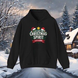 May include: Black hooded sweatshirt with a front graphic that reads "I've Got The Christmas Spirit (It's Gin)". The design includes Santa hats, a star, and a green wreath. The sweatshirt has a front pocket and a drawstring hood.