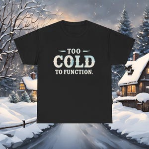 Puede incluir: Camiseta negra con el texto "TOO COLD TO FUNCTION" en blanco y azul claro. La camiseta es de cuello redondo. El fondo muestra una escena invernal con casas y árboles cubiertos de nieve.
