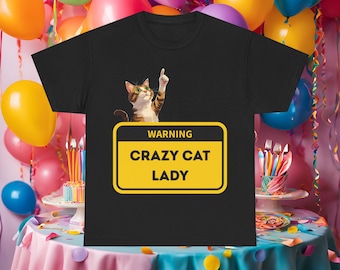 Puede incluir: Camiseta negra con un gato de dibujos animados con gafas señalando hacia arriba. La camiseta tiene un letrero amarillo de advertencia que dice "WARNING CRAZY CAT LADY". El fondo incluye globos coloridos y una mesa decorada.