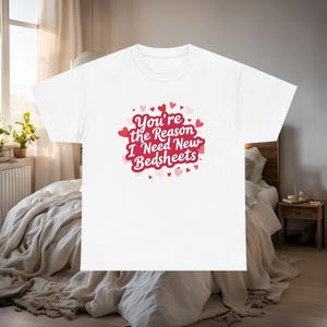 Könnte beinhalten: Weißes T-Shirt mit dem Text "You're the Reason I Need New Bedsheets" in Rot und Rosa, umgeben von Herzformen. Das T-Shirt wird in einem Schlafzimmer gezeigt, was auf ein romantisches oder humorvolles Thema hindeutet.
