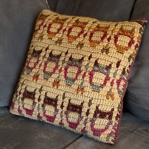 May include: A crocheted pillow with a geometric pattern in shades of brown, pink, orange, and green. The pillow is made with a beige yarn and has a textured surface.