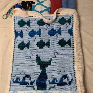 May include: A white crocheted tote bag with a blue and green fish design. The bag has a white crocheted handle and a white crocheted border.