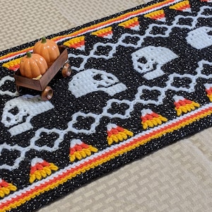 May include: A black and white crocheted table runner with a Halloween theme. The runner features a repeating pattern of candy corn and skulls. The runner has a red, orange, and yellow border. Two small orange pumpkins are sitting on top of the runner.
