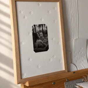 Könnte beinhalten: Ein rechteckiger Bilderrahmen aus hellem Holz mit einem weißen Passepartout, das mit kleinen Sternen geprägt ist. Ein schwarz-weißes Foto einer Person, die ein Kind hält, ist mittig platziert. Der Rahmen steht auf einer Holzstaffelei.