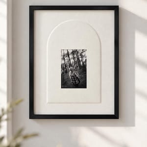 May include: A black rectangular picture frame containing a black and white photograph of a person holding a child in a forest. The photograph is set within a cream-colored mat board featuring an arched cutout design.
