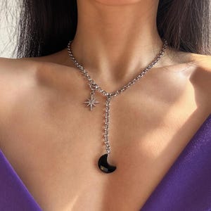 Puede incluir: Un collar de cadena plateada con un dije de media luna negra y un dije de estrella. El collar está en capas y tiene un diseño delicado.
