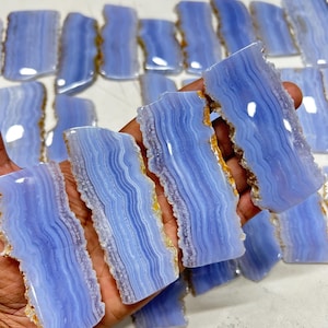 Pode incluir: Uma coleção de placas de ágata rendada azul com uma aparência em camadas e listrada. As placas têm uma cor azul clara com bordas brancas e marrom dourado. As peças de pedra natural têm formato irregular e parecem polidas.