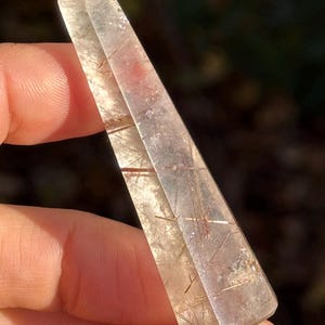 Pode incluir: Uma ponta de cristal de quartzo transparente com inclusões castanho-avermelhadas. O cristal é segurado entre os dedos, mostrando sua estrutura transparente e multifacetada. O cristal é uma pedra preciosa natural e polida.