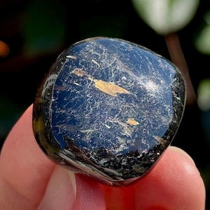 May include: A polished, black gemstone with iridescent inclusions and a smooth, rounded shape. The stone has a reflective surface, catching light and displaying a range of colors. The gemstone is held in a hand, with a blurred green background.