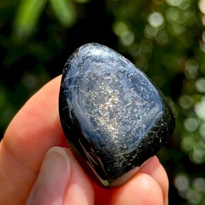 May include: A polished, irregular, black stone with a smooth, glossy surface. The center of the stone features a distinct, shimmering metallic inclusion that reflects light with subtle gold and silver tones, creating a bright, reflective patch.