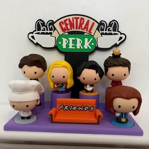 May include: A Friends-themed display featuring miniature figurines of the show's characters. The figurines are arranged on a purple base with an orange couch and the show's logo, "Central Perk". The figurines are approximately 5-7 cm tall.