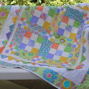 May include: A colorful patchwork quilt with a white background and a variety of floral patterns in shades of green, yellow, purple, and orange. The quilt is folded and draped over a white bench.