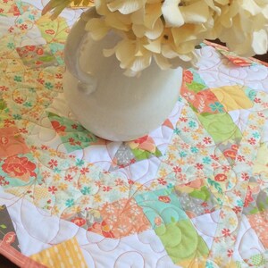 May include: A white quilted table runner with a floral pattern in shades of pink, green, yellow, and blue. The runner is decorated with white stitching in a swirling pattern. A white vase with a bouquet of white flowers is sitting on top of the runner.