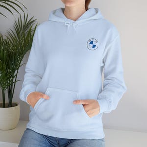 Könnte beinhalten: Hellblauer Kapuzenpullover mit Fronttasche und Kordelzugkapuze. Der Kapuzenpullover hat ein kleines BMW-Logo auf der oberen linken Brust. Das Logo ist ein blau-weißes Emblem in einem schwarzen Kreis.