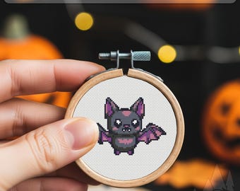 May include: A cross-stitch embroidery hoop featuring a cute cartoon bat with purple wings and a pink heart on its chest. The text "PDF OXS PATTERN" is displayed at the top. The hoop is held in a hand, with pumpkins and decorative lights in the background.