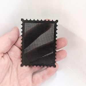 Pode incluir: Um objeto retangular preto com um padrão de grade e bordas recortadas, assemelhando-se à forma de um selo postal. O item apresenta um design de malha aberta consistente em toda a sua superfície, criando uma textura geométrica uniforme.