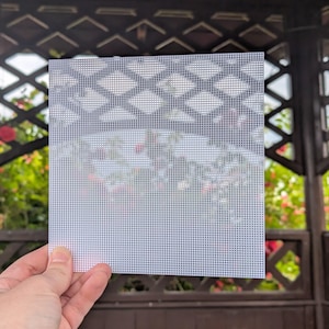 May include: A square sheet of white plastic canvas featuring a uniform grid of small, evenly spaced holes. The material is semi-rigid and semi-transparent, allowing light and the blurred shapes of a garden structure to be seen through the mesh.