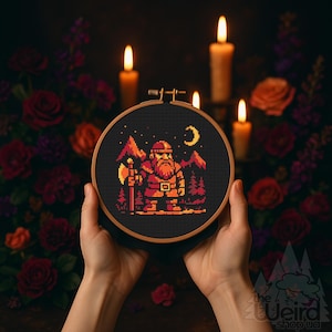May include: A cross-stitch embroidery hoop with a pixelated design of a dwarf with an axe, mountains, and a crescent moon in shades of orange, red, and black. The hoop is held against a dark backdrop with candles and flowers.