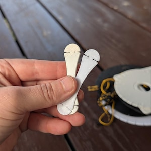 Puede incluir: Dos pequeños organizadores de cables planos de plástico, de forma alargada con extremos redondeados y lados con muescas. Uno es de color blanco roto y el otro gris claro. Ambos tienen una superficie texturizada para sujetar cordones.