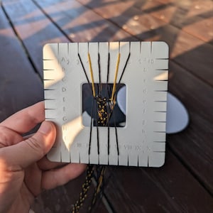 Puede incluir: Un disco de trenzado kumihimo cuadrado de plástico blanco con ranuras numeradas y con letras. Hilos negros y amarillos están enhebrados a través de las ranuras y tejidos en una trenza en la abertura cuadrada central, con un cordón trenzado.
