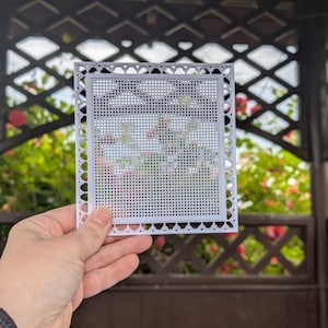 以下が含まれることがあります： 刺繍プロジェクト用の小さな穴のグリッドを備えた正方形の白いプラスチックキャンバスシート。フレームには装飾的なスカラップエッジと円形の切り抜きがあり、刺繍やクロスステッチのデザインのための構造化された境界線を提供します。