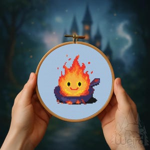 May include: A cross-stitch embroidery hoop featuring a pixelated fire demon with a smiling face. The fire demon is primarily orange and yellow, with a dark blue and purple base. The background is a light blue fabric, and the hoop is held by hands.