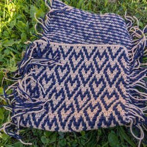Puede incluir: Dos piezas de ganchillo hechas a mano con un patrón geométrico en azul marino y beige. Las piezas tienen flecos en los bordes y están sobre hierba verde. El ganchillo presenta un diseño de chevrón repetido.