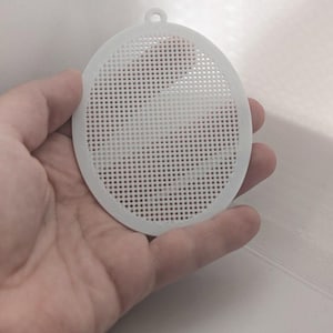 May include: A circular white plastic mesh disc featuring a uniform grid pattern of small square perforations throughout its surface. The disc includes a small integrated loop at the top edge, designed for hanging or attachment purposes.