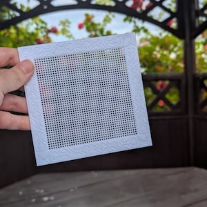 Peut inclure: Une feuille de canevas en plastique blanc de forme carrée, dotée d'une grille de petits trous pour la broderie. Le canevas est entouré d'une bordure décorative avec des motifs en relief de volutes et de cœurs sur les quatre côtés.