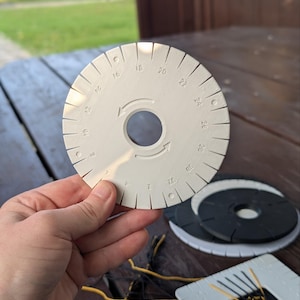 Puede incluir: Un disco de kumihimo circular de plástico blanco con un orificio central, ranuras numeradas del 2 al 32 y flechas direccionales. El disco presenta muescas espaciadas uniformemente a lo largo del borde exterior para enhebrar cordones.