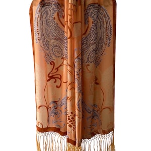 May include: Peach-coloured scarf featuring a paisley design in shades of purple, brown, and white. The scarf has a fringe trim in a matching peach colour. The scarf is draped, displaying the intricate pattern and texture.