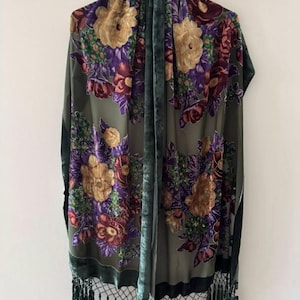 Op de afbeelding: Olijfgroene bloemen sjaal met een fluwelen textuur. De sjaal heeft grote, gedetailleerde bloemen in tinten goud, paars en bordeaux. De randen zijn afgewerkt met een donkergroene rand en zwarte franjes.