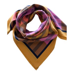 May include: A square silk scarf featuring a bold plaid pattern in shades of mustard yellow, pink, brown, and navy blue. The scarf is folded to show a central check design with a solid mustard yellow border and a thin navy blue inner stripe.