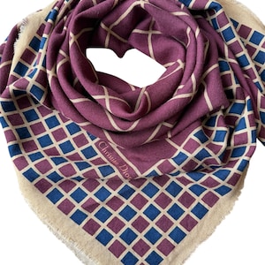 May include: A square scarf with a geometric pattern in shades of purple, blue, and beige. The scarf features a repeating diamond design and a fringed edge. The text "Christian Dior" is visible on the scarf.