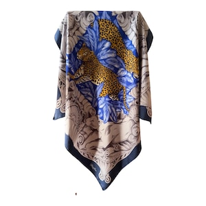 May include: A square scarf featuring a central blue panel with two leopards surrounded by blue foliage. The border displays ornate grey and white scrollwork patterns on a dark navy background. The fabric has a smooth finish with a visible brand logo.