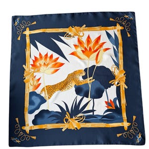 Puede incluir: Pañuelo cuadrado de seda con borde azul marino y un diseño central con un leopardo saltando, flores de loto naranjas y follaje azul. El diseño está enmarcado por un borde estilo bambú con detalles dorados.