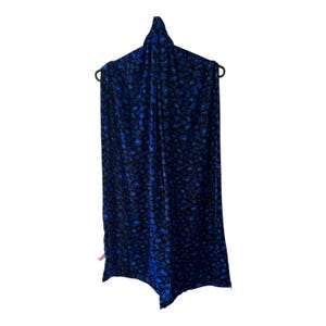 Puede incluir: Una bufanda rectangular con un vibrante estampado animal en azul y negro. La tela tiene una textura suave similar al terciopelo y está colgada en una percha negra. El diseño consiste en formas irregulares y redondeadas en azul brillante sobre un fondo negro oscuro, creando un efecto visual audaz y de alto contraste en toda la superficie de la prenda.