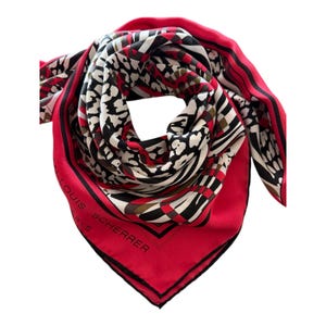 May include: A square scarf featuring a vibrant red border with the text Jean-Louis Scherrer Paris printed in black. The center displays a bold graphic pattern of black, white, and red abstract shapes, including leopard-like spots and geometric lines.