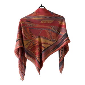 Peut inclure: Un foulard carré orné d'un motif géométrique dans les tons de rouge, orange et marron. Il est suspendu à un cintre noir sur fond blanc. Le motif comprend des formes et des lignes abstraites.