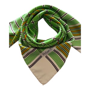 Puede incluir: Un pañuelo cuadrado con un vibrante patrón de rayas y lunares en verde, blanco y naranja con detalles en marrón oscuro. La tela muestra el texto Yves Saint Laurent en letras negras cerca de la esquina sobre un fondo de color crema sólido.