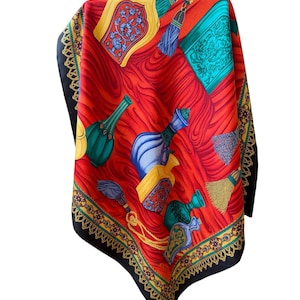 May include: A silk scarf with a vibrant red background featuring a pattern of ornate perfume bottles in teal, yellow, and blue. The scarf has a black border with a gold and green decorative trim and tassels.