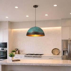 Peut inclure: Un luminaire suspendu turquoise et bronze dans une cuisine. Le luminaire en forme de dôme a un extérieur turquoise texturé, une bande de bronze et un intérieur doré. La cuisine comprend des armoires blanches, un réfrigérateur en acier inoxydable et un comptoir en marbre.