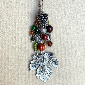 May include: A silver-toned charm with a leaf pendant and a cluster of beads in shades of green, orange, and brown. The charm includes a pinecone and a lobster clasp for attachment. The beads are various shapes and sizes.