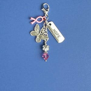 Op de afbeelding: Een zilveren bedel met een karabijnsluiting, met een roze lint, een vlinder, een bloem, een roze kristal en een label met de tekst "HOPE". De bedel is ontworpen om aan een armband of ketting te worden bevestigd.