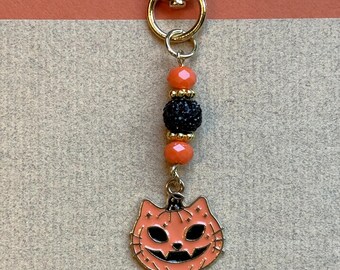 May include: A Halloween-themed keychain with a gold-tone clasp and hardware. The charm features a smiling jack-o'-lantern face in orange and black enamel. The keychain also includes orange and black beaded accents.