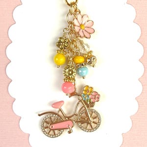 May include: A gold-tone keychain charm featuring a pink bicycle with a basket of flowers, pink and yellow beads, and a pink flower. The charm is attached to a gold heart-shaped clasp.
