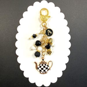 May include: A gold-toned keychain with a teapot charm, black and white beads, and a black letter "A" charm. The teapot has a black and white checkered pattern. The keychain is attached to a gold-colored clasp.